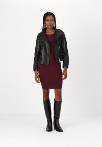 Crna kožna jahačka jakna s preklopom, uparena s burgundskom čipkastom haljinom i crnim kožnim čizmama do koljena.