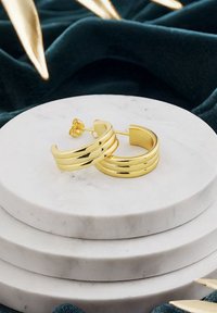Orecchini a cerchio in oro con un design a triplo anello, caratterizzati da una finitura liscia e lucida. Esibiti su un piedistallo in marmo impilato.