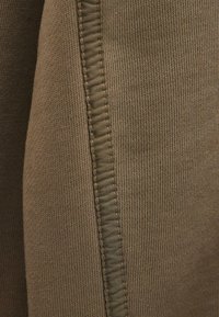 Bruine geaccentueerde stof met zichtbare ribbels en een contrasterende gladde naaddetail. Het materiaal lijkt zacht en flexibel.