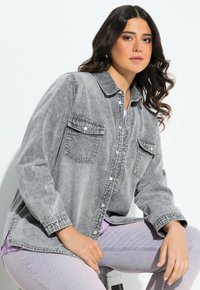 Camicia di denim grigia con maniche lunghe, due tasche sul petto, bottoni frontali e dettagli cuciti, indossata sopra un top viola e jeans chiari.