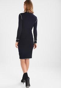 Granatowa sukienka swetrowa z prążkowanej dzianiny, z długimi rękawami, dopasowaną sylwetką i białymi falbaniastymi akcentami przy mankietach i u dołu. Noszona z czarnymi botkami na obcasie.