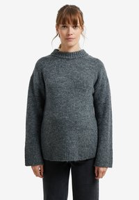 Frau mit hellbraunem Haar, die einen dicken grauen gestrickten Pullover und schwarze Hosen trägt, steht vor einem schlichten weißen Hintergrund.