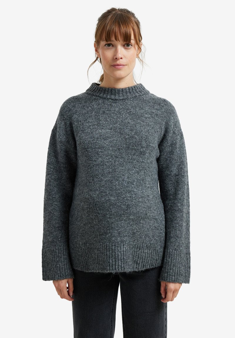 Frau mit hellbraunem Haar, die einen dicken grauen gestrickten Pullover und schwarze Hosen trägt, steht vor einem schlichten weißen Hintergrund.
