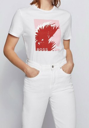 Femme portant un t-shirt à manches courtes blanc avec imprimé abstrait de feuilles rouges et un jean blanc taille haute, mains dans les poches, fond neutre.