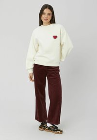 Sweatshirt blanc avec un graphique de cœur rouge et un design en rayon de soleil, associé à un pantalon en velours côtelé bordeaux. Des chaussures noires et beiges à motif complètent le look.