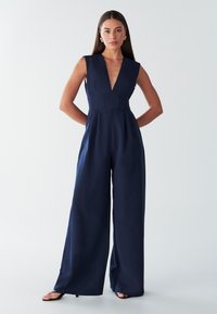 Temno modra kombinezon z globokim V-izrezom in brez rokavov. Ponaša se z oprijetim zgornjim delom, širokimi hlačnicami in gladkim, teksturiranim materialom.