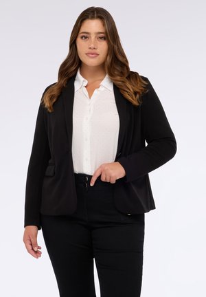 Blazer nero con vestibilità su misura, texture morbida e due tasche frontali, indossato sopra una blusa bianca a pois neri e pantaloni neri.