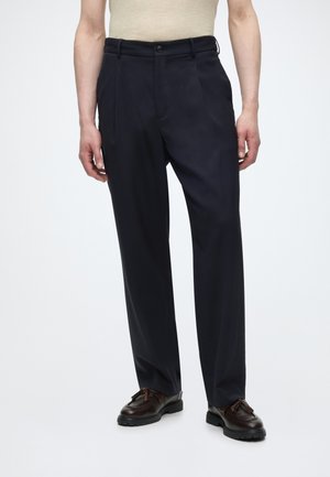 Férfi fekete, pliszírozott, szabott nadrággal és fekete bőr bojtos cipővel, sima fehér háttér előtt áll.