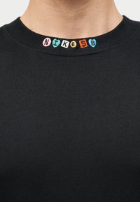 Μαύρο μπλουζάκι με στρογγυλή λαιμόκοψη με πολύχρωμα κεντημένα γράμματα που σχηματίζουν τη λέξη "NIKE SB" κατά μήκος της λαιμόκοψης στο πάνω μέρος του στήθους ενός ατόμου.