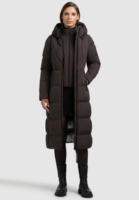 Lång puffjacka i mörkbrun med kviltad design, huva, framsidans dragkedja och sidofickor. Strukturerad tyg med slät innermaterial.