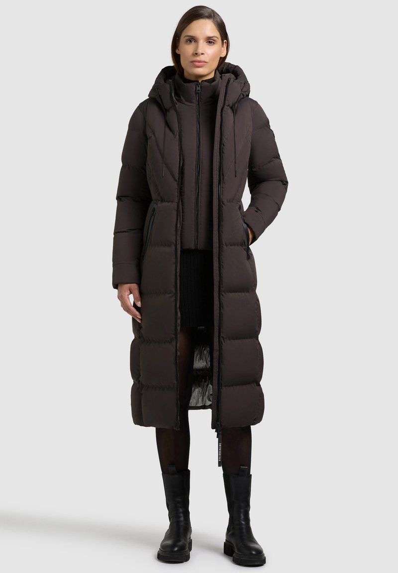 Lång puffjacka i mörkbrun med kviltad design, huva, framsidans dragkedja och sidofickor. Strukturerad tyg med slät innermaterial.