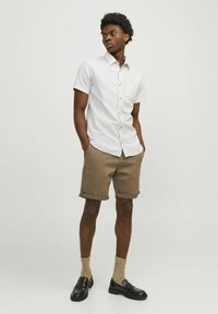 Camisa blanca de manga corta con botones y un bolsillo, combinada con pantalones cortos marrones arremangados. Mocasos negros y calcetines beige completan el atuendo.
