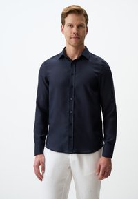 Marineblauwe langemouwen shirt met een klassieke kraag, knoopsluiting aan de voorkant en een gladde textuur; gedragen met lichtgekleurde broek.