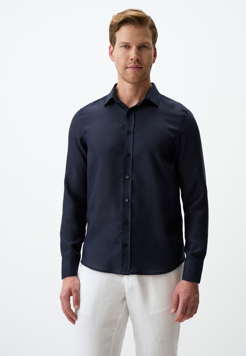 Marineblauwe langemouwen shirt met een klassieke kraag, knoopsluiting aan de voorkant en een gladde textuur; gedragen met lichtgekleurde broek.