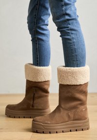 Bruna mockaskor vid ankeln med en beigefärgad fuskpälskrage, texturerad sidodragkedja och en tjock gummisula med ett zigzagmönster.