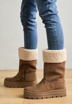 Brązowe zamszowe botki za kostkę z beżowym futerkiem, teksturowanym zamkiem z boku oraz grubą gumową podeszwą w wzór zygzaka.