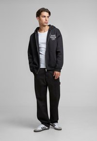 Felpa nera con zip con la scritta "Sunday Running Club" in bianco, abbinata a una canotta bianca e pantaloni neri larghi, completata da sneakers bianche.