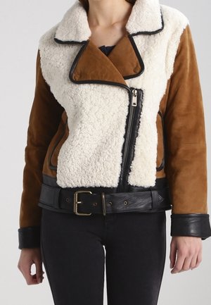 Woman wearing brown suede and cream shearling jacket with black leather belt, cuffs, and an asymmetrical front zipper, paired with black pants.