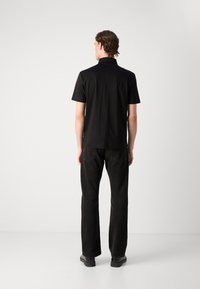 Black short-sleeve polo shirt with a collar, made of smooth fabric. Worn with dark pants, viewed from the back against a plain background.