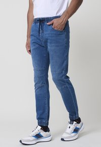 Pantalones jogger de mezclilla azul con cintura elástica, dobladillos ajustados, bolsillos laterales y detalles en panel en los muslos. Zapatillas blancas visibles.