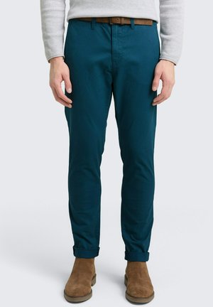 Mies, joka käyttää teal-värisiä slim-fit-housuja, joissa on käänteet, ruskeaa vyötä, beigeä neuletta ja ruskeita mokka nilkkabootseja, seisoo neutraalia taustaa vasten.