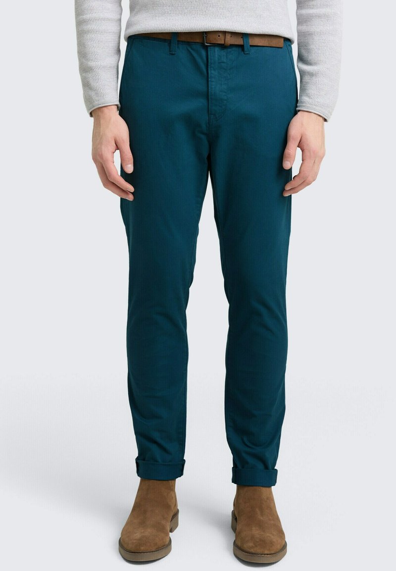 Muškarac u tirkiznim slim-fit hlačama s podvrnutim nogavicama, smešnim pojasom, bež džemperom i smešnim nubuk čizmicama stoji naslonjen na neutralnu pozadinu.