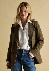 Pruunist plaaditud bleiser, millel on struktureeritud lõige ja kuldsed nupud, millele on lisatud valge nööbiga särgi ja kõrge vöökohtadega siniste teksadega.