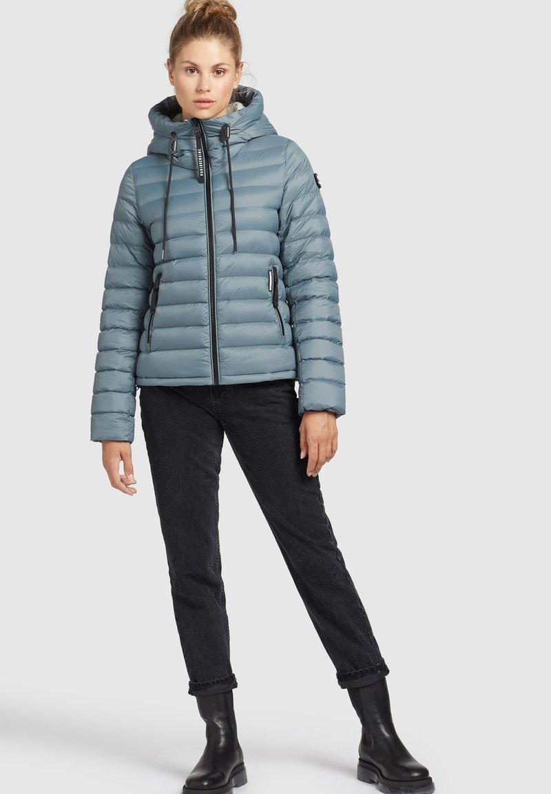 Gepolsterte, hellblaue Jacke mit Kapuze, mit vertikalem Steppmuster und schwarzen Reißverschlüssen. Kombiniert mit dunklen Jeans und schwarzen Stiefeln.