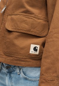 Geacă Carhartt maro cu buzunar mare cu clapetă și logo vizibil, asortată cu blugi albaștri la talie.
