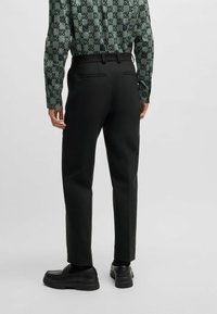 Pantaloni neri con vestibilità sartoriale, dotati di due tasche posteriori, abbinati a una camicia verde a maniche lunghe con motivo e scarpe nere senza lacci.