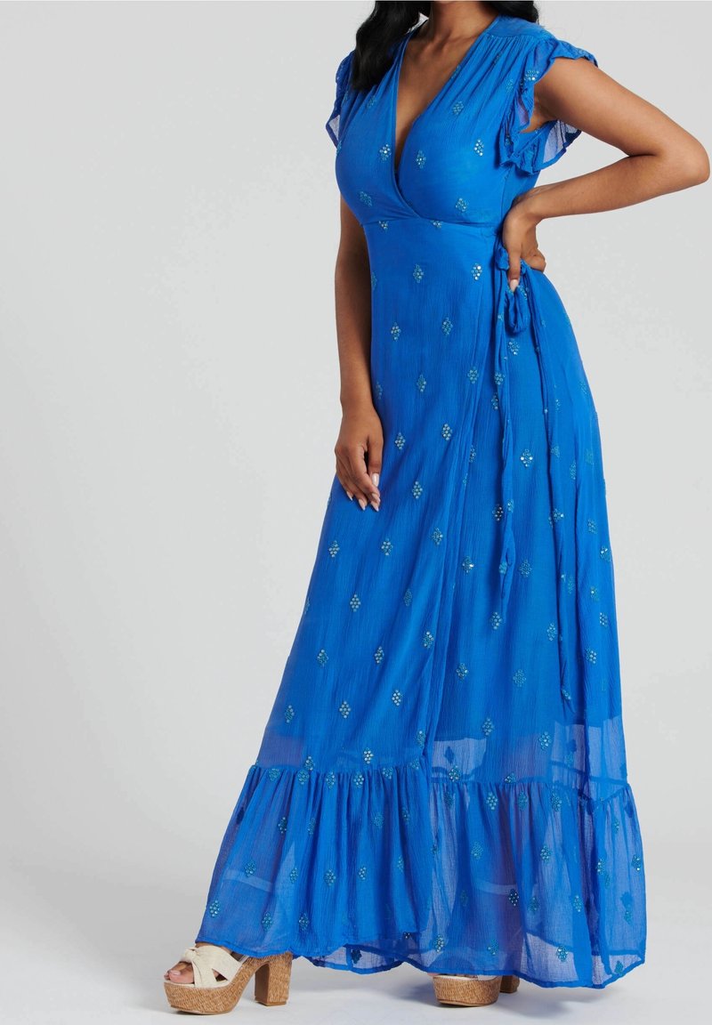 Femme portant une robe portefeuille bleue longue jusqu'au sol avec des manches volantées, des embellissements argentés et des talons compensés beiges, debout, une main sur la hanche.