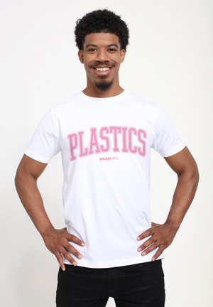 Homme souriant aux cheveux bouclés portant un t-shirt blanc avec un grand texte rose "PLASTICS" et un plus petit "MEAN GIRLS", mains sur les hanches.