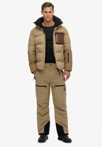 Tan farvet pufferjakke med en brun brystlomme, kombineret med matchende tan bukser med sorte detaljer og lynlåslommer, båret med sorte støvler.