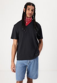 Zwarte T-shirt met korte mouwen en een relaxte fit, gecombineerd met lichtblauwe denimshorts. Een rode bandana is om de nek gedragen.