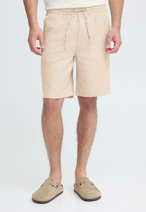Beige Leinen-Shorts mit elastischem Bund und Kordelzug. Enden auf halber Oberschenkellänge mit lockerem Sitz. Kombiniert mit beigen Slippern.
