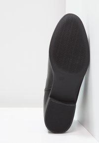 Zapato negro con una parte superior de cuero liso y una suela de goma texturizada con patrones de olas. Tamaño marcado como 37 en la suela.