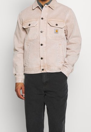 Mand iført en lysebeige skjortejakke med brystlommer og et lille logoemblem, kombineret med mørke bukser og en skjorte med krave.