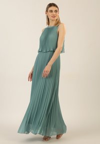 Türkisfarbene, plissierte Maxikleid mit schulterfreiem Design, fließendem Stoff und geschichtetem Oberteil. Texturierte Falten verstärken die visuelle Tiefe.