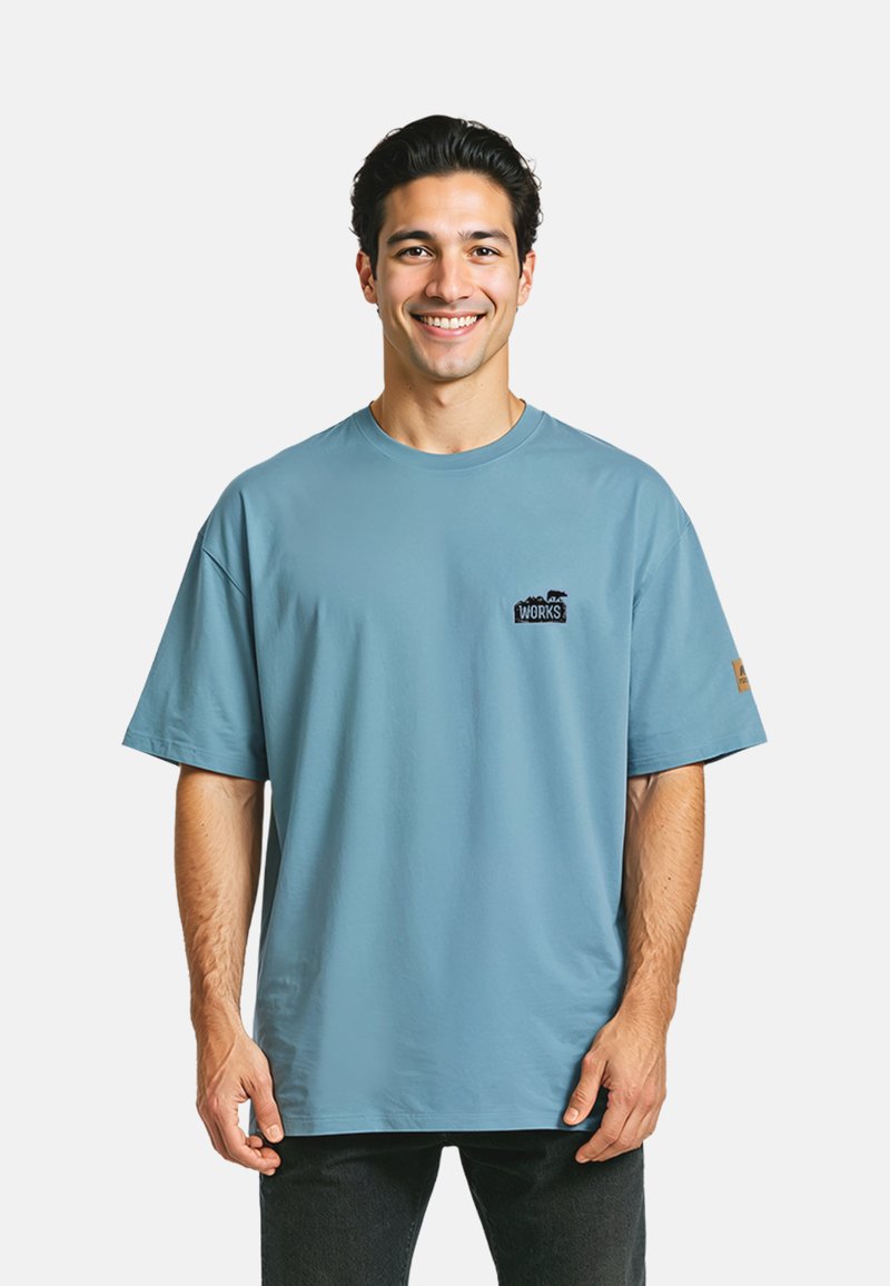 Lichtblauwe katoenen t-shirt met korte mouwen en een ronde hals, voorzien van een klein zwart logo op de borst en een stoffen patch op de mouw.