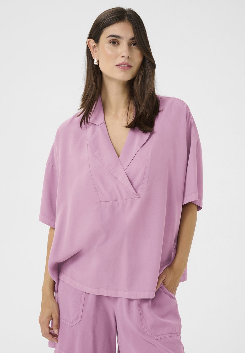 Lillā oversize blūze ar V-veida kakla izgriezumu, īsām piedurknēm un brīvu piegriezumu. Gluda tekstūra un minimālistisks dizains. Saskaņota ar atbilstošām šortiem.