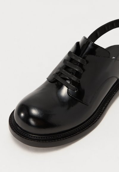 Black patent leather shoe with a rounded toe, laced front, and open back. Features a textured sole and smooth, glossy finish.