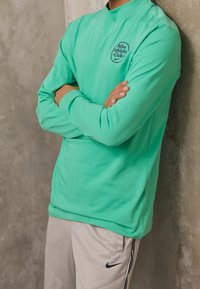 Light green Nike Athletic Club sweatshirt with long sleeves and a high neckline, paired with gray pants featuring a black logo.
