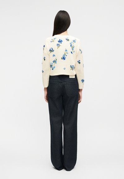 Γυναίκα με μακριά σκούρα μαλλιά που φοράει κρεμ πουλόβερ πλεκτό με μπλε ανθισμένα μοτίβα και χαλαρό μαύρο παντελόνι, στέκεται γυρισμένη με την πλάτη σε απλό φόντο.