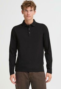 Čierna polokošeľa s dlhým rukávom a golierom, tromi gombíkmi a textúrovaným povrchom, v kombinácii s hnedými nohavicami.