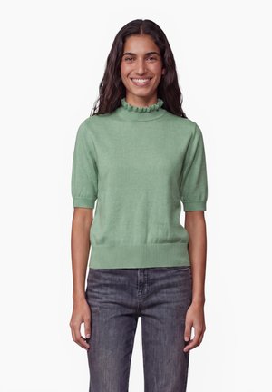 Femme souriante aux longs cheveux foncés portant un pull vert clair à manches courtes avec col à volants et un jean gris foncé, debout devant un fond blanc.