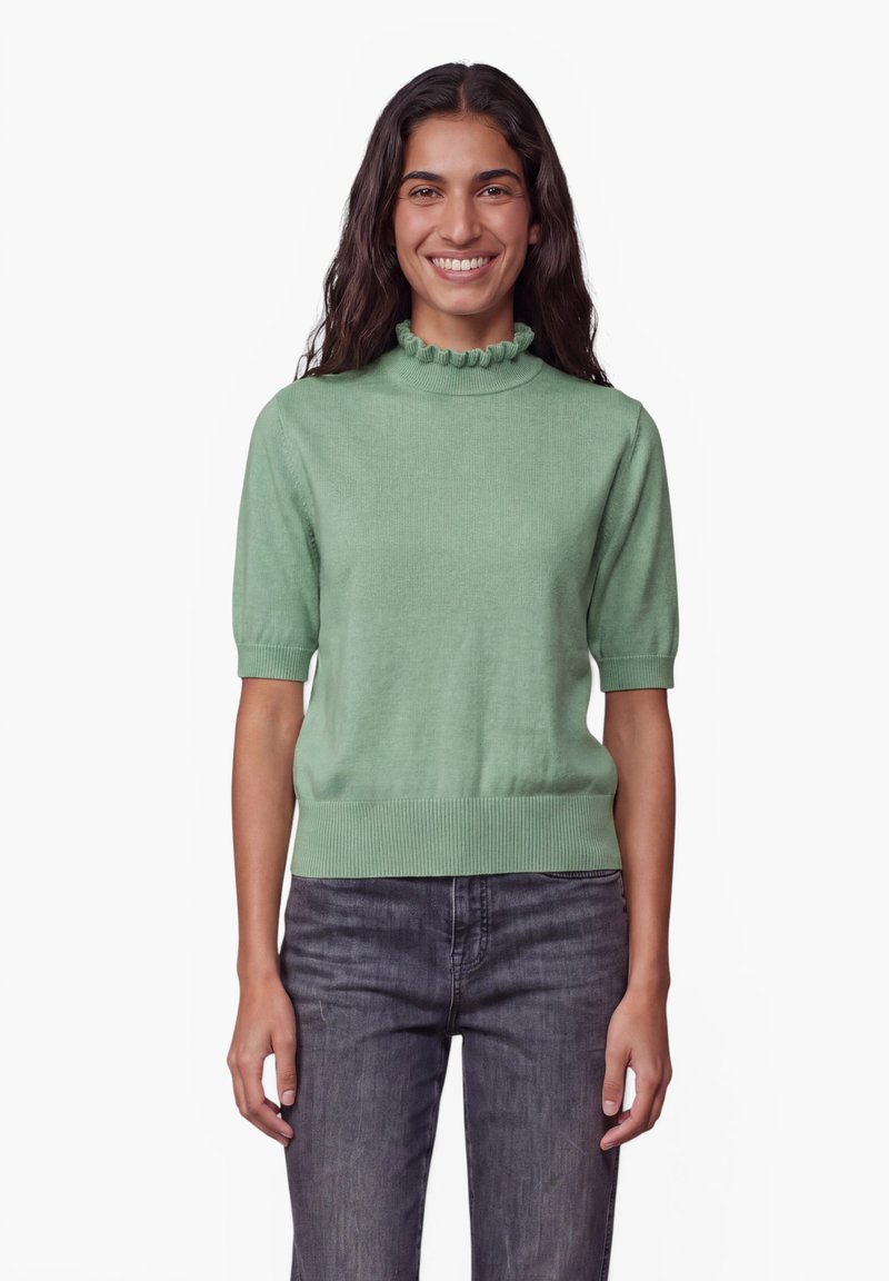 Femme souriante aux longs cheveux foncés portant un pull vert clair à manches courtes avec col à volants et un jean gris foncé, debout devant un fond blanc.