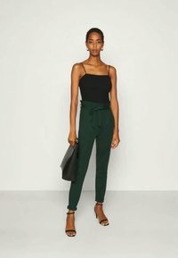 Top noir sans manches, pantalon vert ceinturé, sac à main noir, talons avec lanières, tissu lisse, coupe ajustée, assis à la taille, matériel minimal.