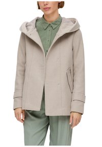 Femme portant un manteau en laine beige à capuche sur une chemise verte à boutons et un pantalon vert assorti, montrée du cou à mi-cuisse, debout.