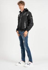 Giacca a vento nera con dettagli lucidi, tasche con zip, polsini elasticizzati e orlo regolabile, abbinata a jeans blu strappati e sneakers bianche.