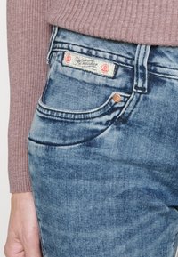 Světle modré džínové kalhoty s přední kapsou, vybledlou texturou a nášivkou s červenými akcenty a logem přišitým v pase.